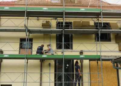 Fassade eines umgebauten Gebäudes mit Gerüstaufbau: Mehrere Arbeiter verrichten an der Außenwand Sanierungsarbeiten, während die Bauleitung den Fortschritt koordiniert.